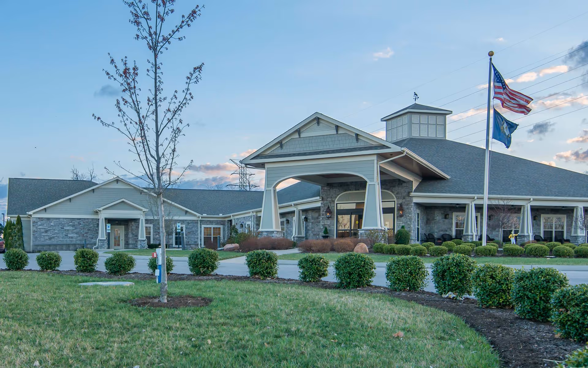 Exterior view of Morning Pointe of Lexington facility with a large covered entrance, stone and siding facade, manicured bushes, a small tree in the foreground, and two flagpoles displaying the American flag and another flag against a partly cloudy sky.