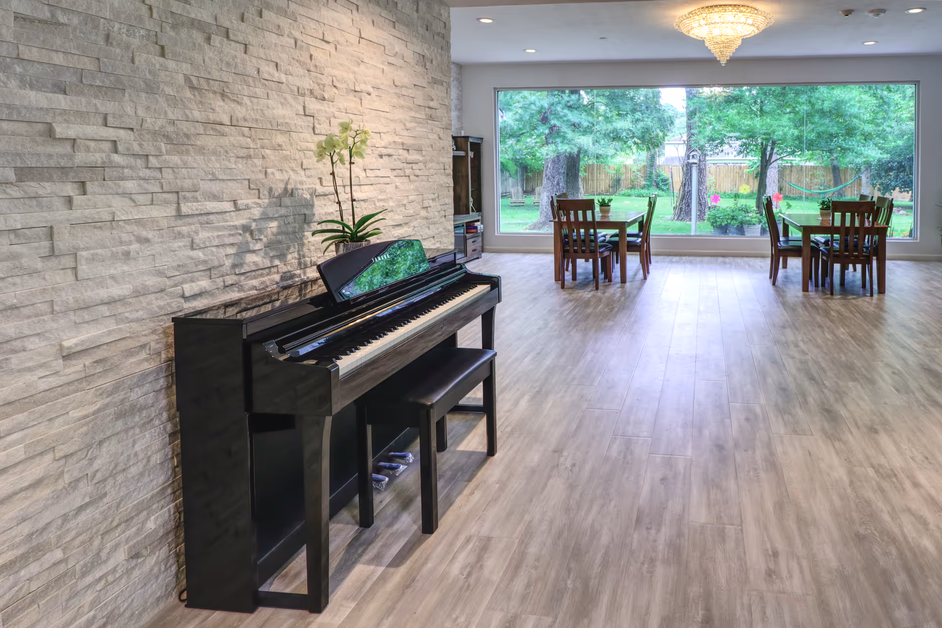 A spacious room with a black upright piano against a textured stone wall on the left. Two wooden tables with chairs are placed near a large window that looks out onto a green garden with trees and a hammock. A chandelier hangs from the ceiling, and the floor is covered with light wood laminate.
