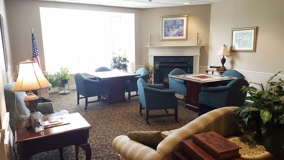 Cozy common room with upholstered chairs and tables around a fireplace and a large window.