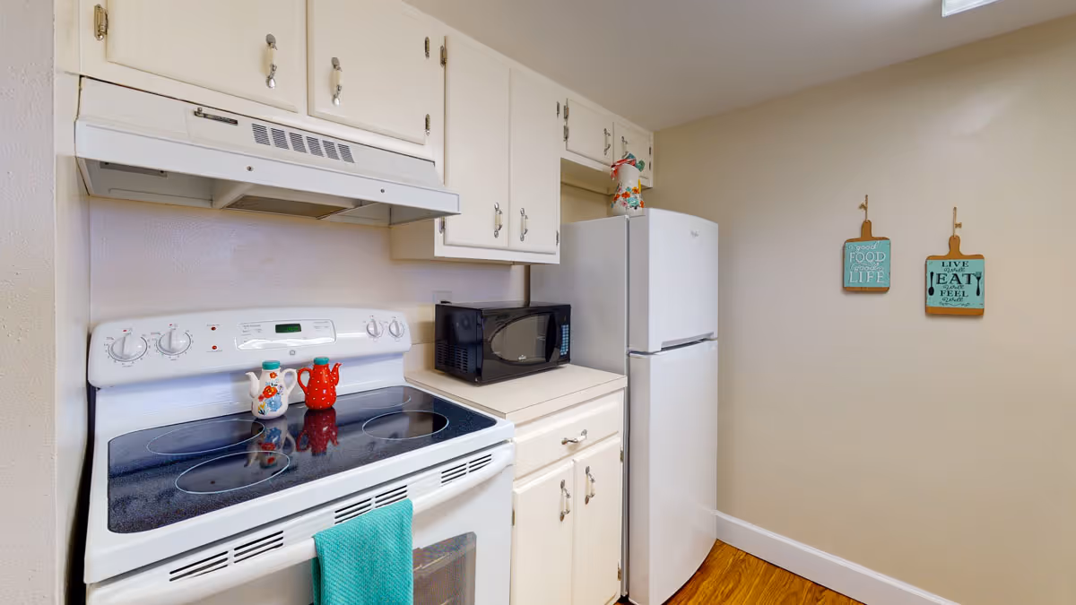 A small kitchen area with a white electric stove and oven, a black microwave on the countertop, and a white refrigerator. There are white cabinets above and below the countertop. Two decorative wall hangings with food-related quotes are on the beige wall. The floor is wooden.