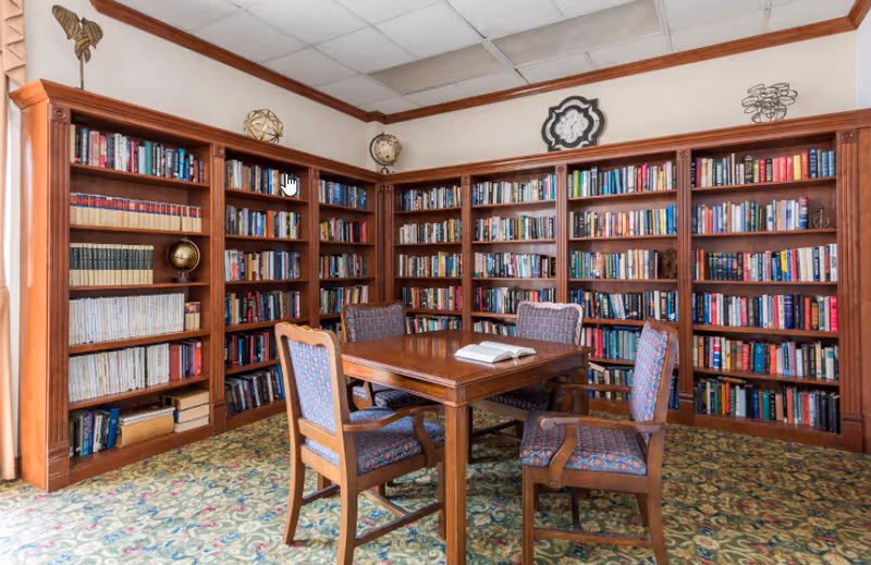 A cozy library room with wooden bookshelves filled with books lining the walls. In the center, there is a wooden table with four upholstered chairs around it. An open book lies on the table. The room has a patterned carpet and decorative items on top of the shelves, including a globe and a clock on the wall.