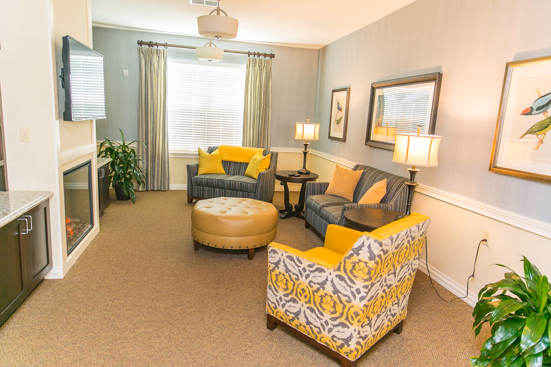 A cozy living room with a striped loveseat and sofa, both adorned with yellow and beige cushions. A patterned armchair with yellow accents faces a round, tufted ottoman in the center. Two table lamps on dark wooden side tables provide warm lighting. A flat-screen TV is mounted above a modern electric fireplace on the left wall. The room has beige carpeting, light-colored walls with a chair rail, framed bird artwork, and a large window with striped curtains letting in natural light. There are also two green potted plants adding a touch of nature to the space.