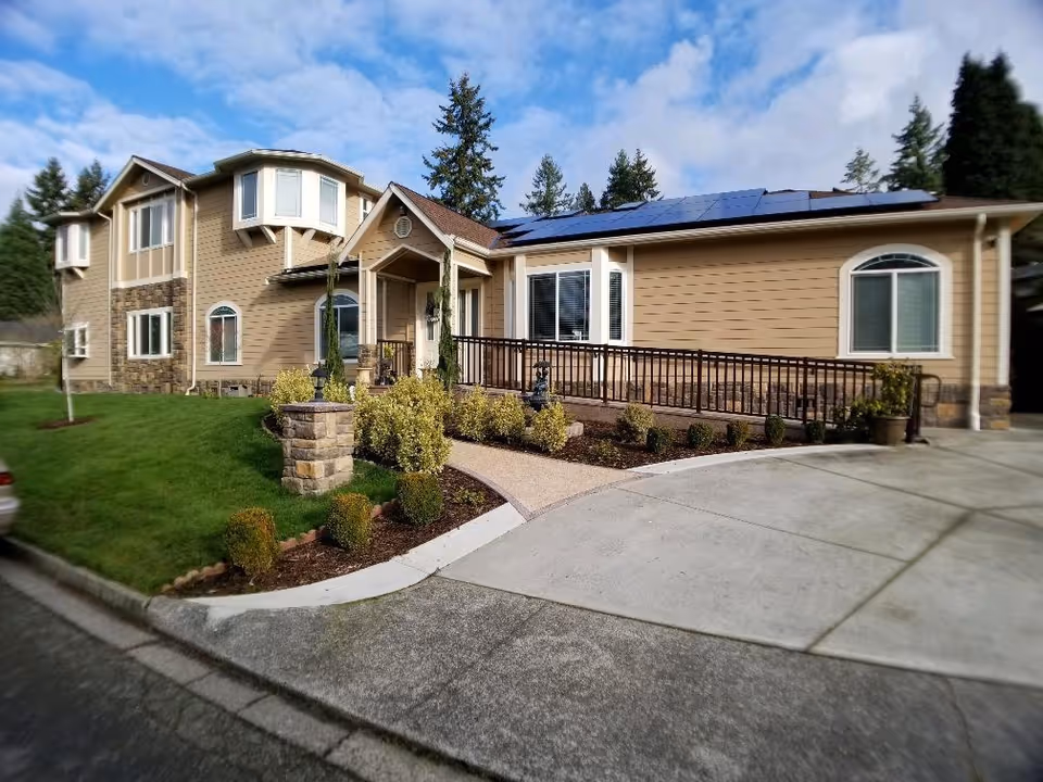 Exterior view of a two-story senior care facility building with beige siding, stone accents, and solar panels on the roof. The entrance has a small covered porch with a ramp and landscaped bushes along the walkway. The sky is partly cloudy and there are tall trees in the background.