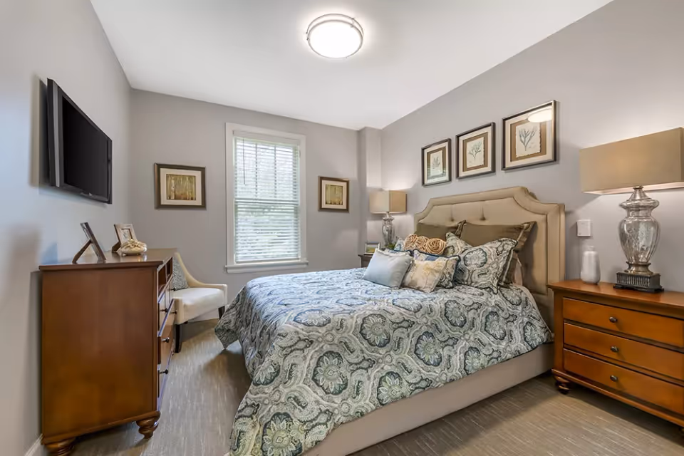 A cozy bedroom with a large bed covered in a patterned blue and gray comforter and multiple pillows. The room has light gray walls, a window with blinds, a wall-mounted flat-screen TV, a wooden dresser, a white upholstered chair, two wooden nightstands with lamps, and framed artwork on the walls.