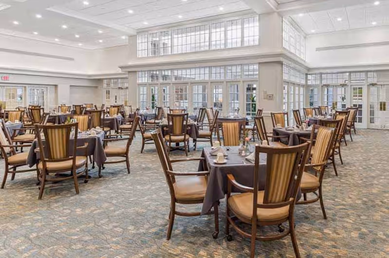 A spacious dining room with multiple tables covered in dark tablecloths, each surrounded by wooden chairs with cushioned seats and backs. The room has large windows allowing natural light to fill the space, and the ceiling is high with recessed lighting.