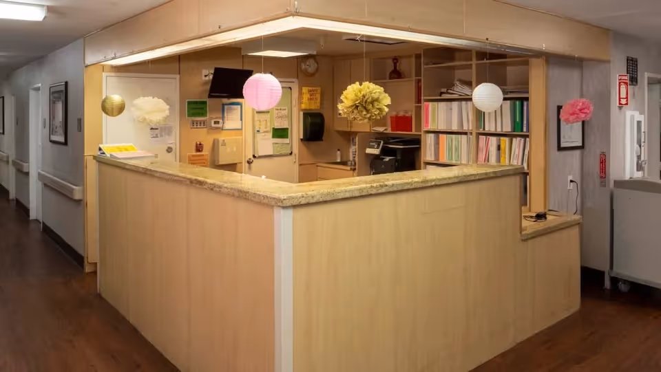 Reception desk area inside a healthcare facility with hanging paper lantern decorations, shelves filled with binders, a printer, and various notices on the walls. The area has wood flooring and light-colored wood paneling on the desk and walls.