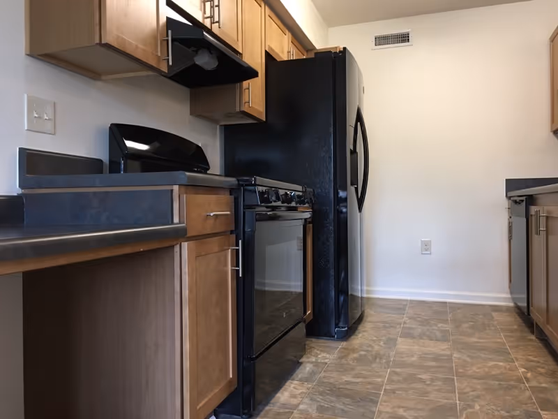 A kitchen with wooden cabinets, black appliances including a stove, oven, and refrigerator, and a tiled floor. The walls are plain white with a vent and electrical outlet visible.