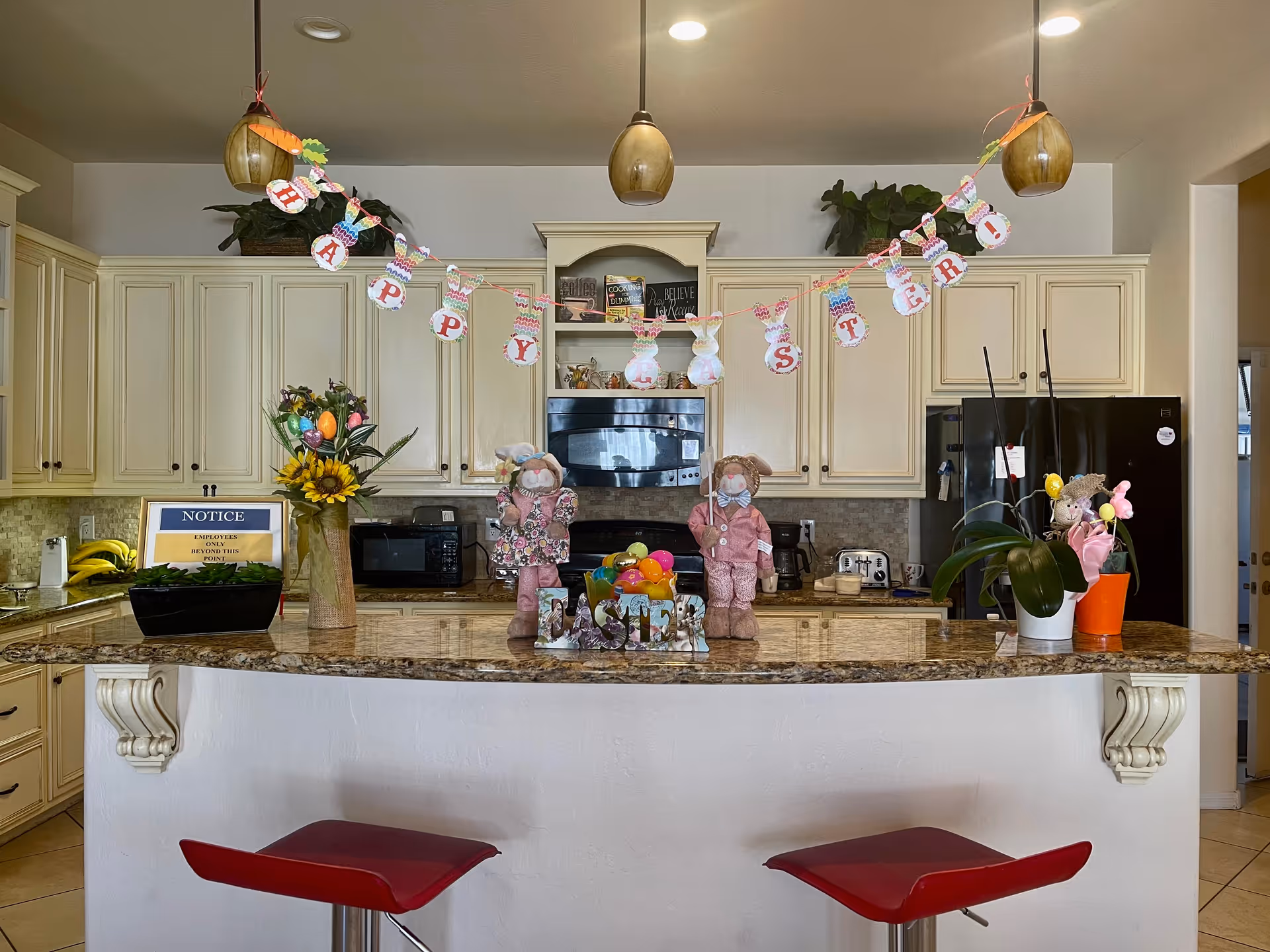 Bright kitchen with a marble-topped island decorated for Easter with a banner, dolls, flowers and two red bar stools.