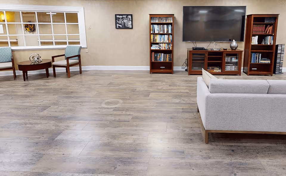 A spacious living room area with light wood flooring, a light gray sofa on the right, two wooden bookshelves filled with books, a large flat-screen TV mounted on the wall above a wooden TV stand, and two armchairs with a round wooden table between them on the left side. A black and white framed photo is hung on the beige wall.