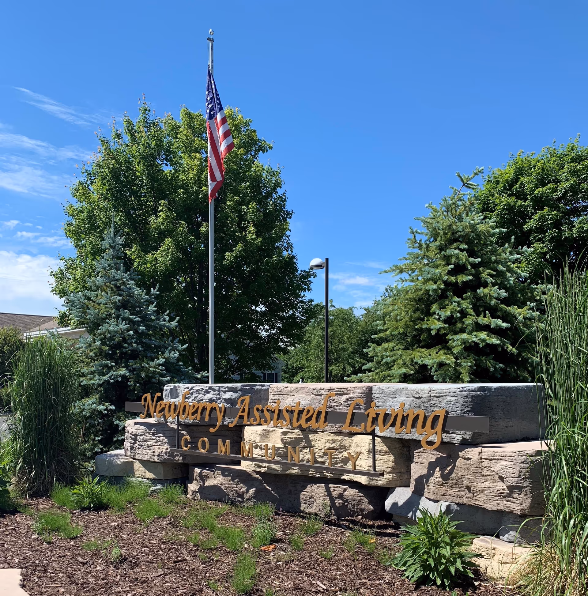 Outdoor view of the Newberry Assisted Living Community sign made of stone with gold lettering, surrounded by green trees, plants, and a flagpole with an American flag against a clear blue sky.