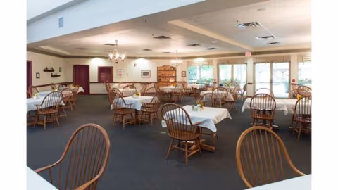 A spacious dining room with multiple wooden tables and chairs arranged neatly. Each table is covered with a white tablecloth and has a small centerpiece. The room has large windows allowing natural light to enter, and there are chandeliers hanging from the ceiling. The walls are light-colored with some framed pictures and shelves.