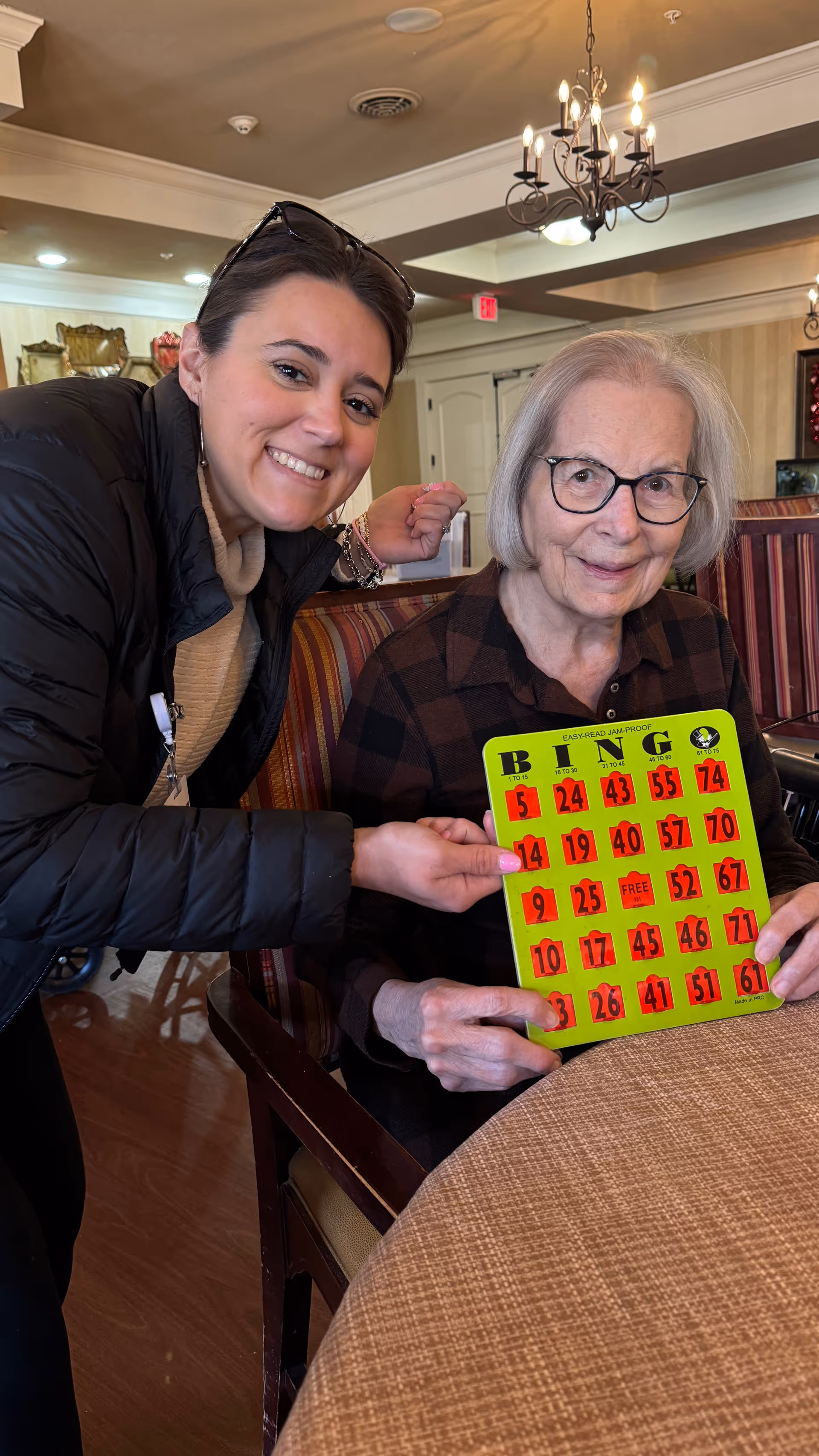Two smiling women in a senior living dining area holding a bright green bingo card.