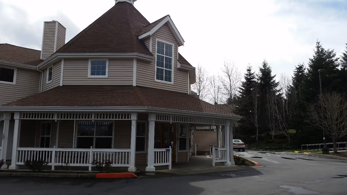 Exterior view of a building with beige siding and a brown roof, featuring a covered porch with white railings and columns. There are trees and a parked car in the background under a cloudy sky.