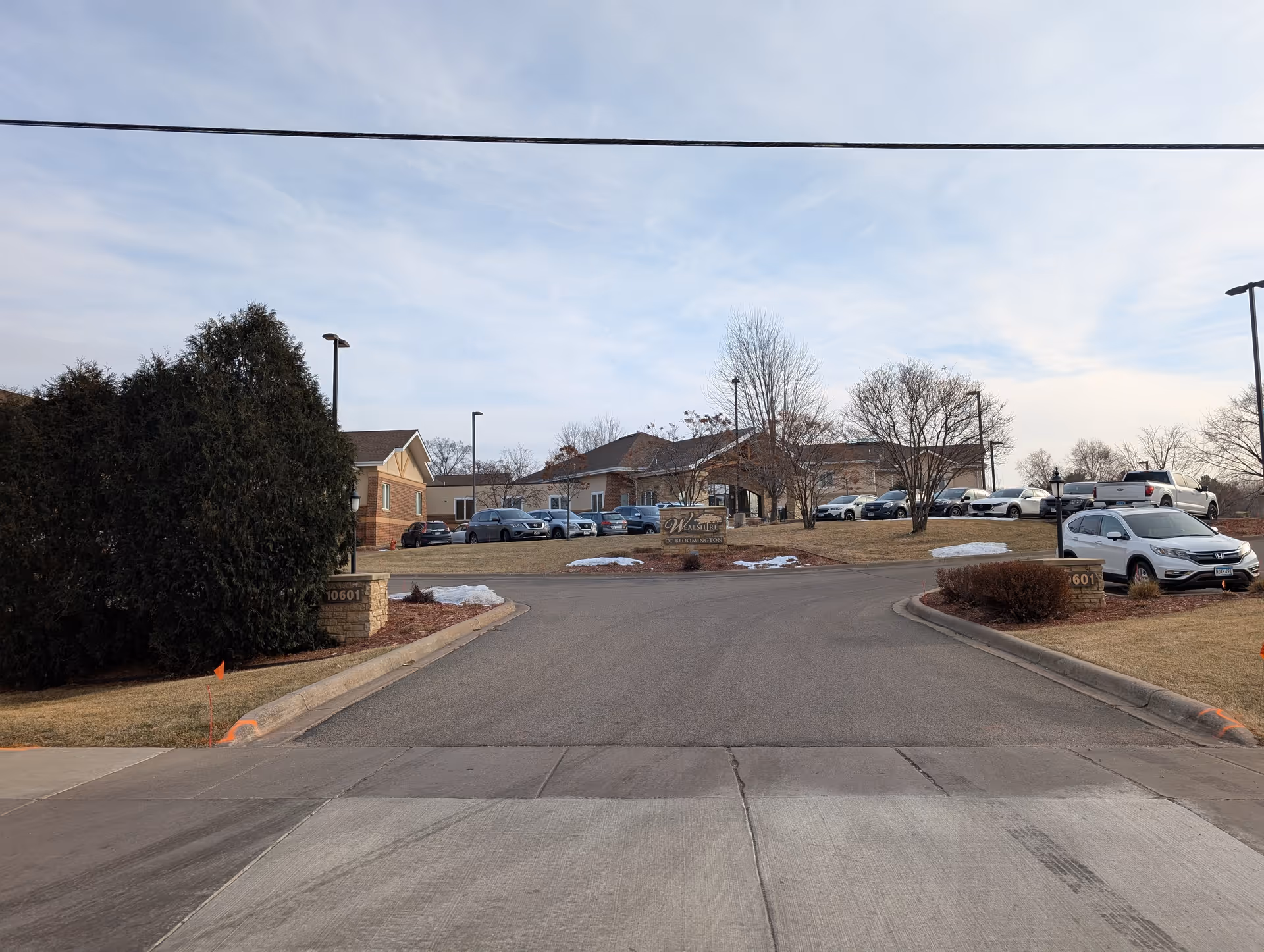 View of the entrance driveway to Wealshire of Bloomington senior living facility, showing the building in the background, parked cars, some leafless trees, patches of snow on the grass, and a sign with the facility's name.