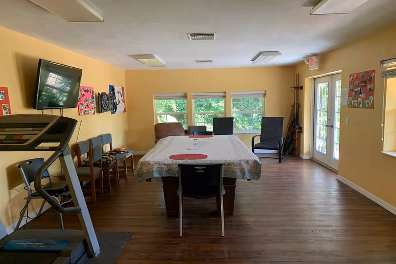 A room with yellow walls and wooden flooring featuring a table covered with a white and floral tablecloth in the center, surrounded by chairs. There is a treadmill on the left side, a wall-mounted TV above a row of chairs, and windows with greenery visible outside. On the right side, there are glass doors leading outside and colorful posters on the walls.