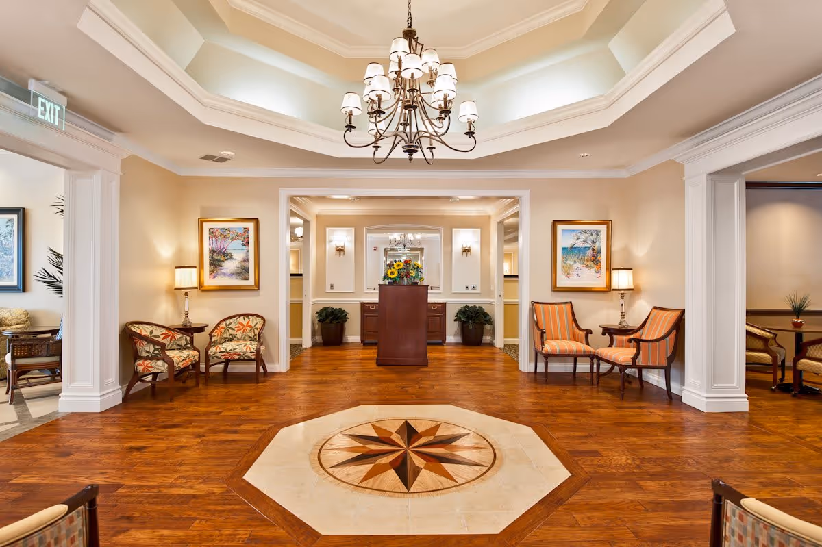 A spacious and elegant interior common area with wooden flooring and a decorative compass design in the center. The room features a large chandelier hanging from a tray ceiling, framed paintings on the walls, and several upholstered chairs arranged in pairs with small tables and lamps. There are potted plants and a wooden podium or stand with a flower arrangement in the background.
