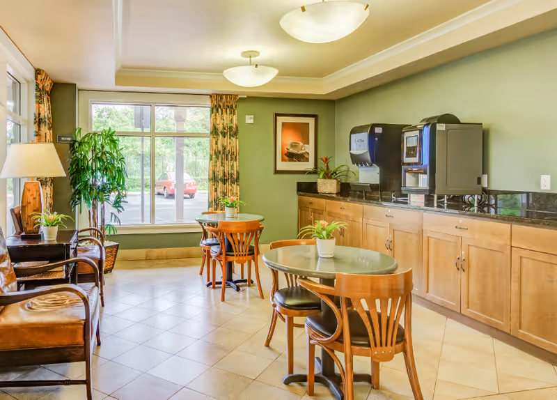 A bright and clean common area with green walls and tiled floor featuring round tables with wooden chairs, small potted plants on the tables, a coffee and water dispenser on a wooden cabinet, a large window with patterned curtains, a floor lamp, and a framed picture on the wall.