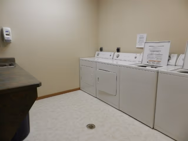 Laundry room with three white washing machines and one dryer lined up against a beige wall. There is a countertop with a sink on the left side and a soap dispenser mounted on the wall above it. The floor is light-colored with a drain in the center.