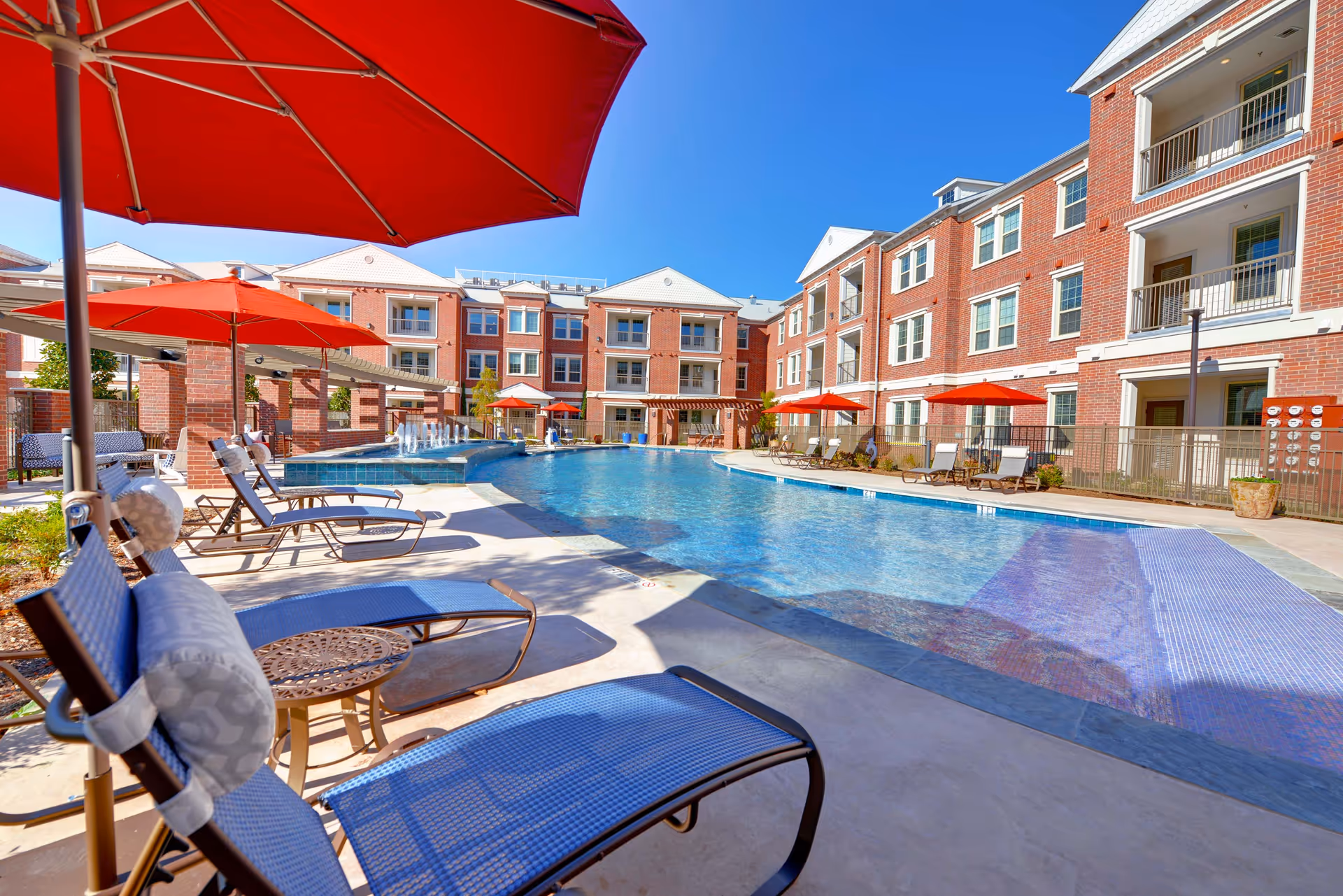 Courtyard pool area with lounge chairs and red umbrellas framed by a three-story brick senior living building under a clear blue sky.