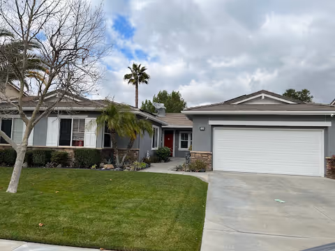 Front exterior view of a single-story residential building with a gray facade, white garage door, red front door, and a well-maintained lawn with some trees and shrubs under a partly cloudy sky.