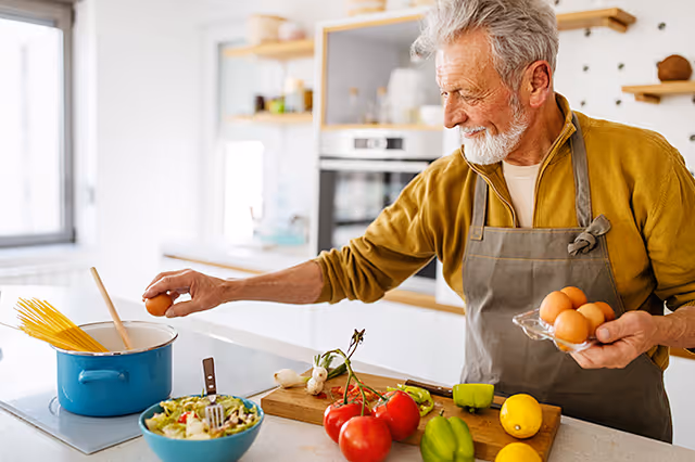 An elderly man wearing a gray apron and mustard yellow shirt is cooking in a modern kitchen. He is holding a carton of eggs in one hand and cracking an egg into a blue pot on the stove. Fresh vegetables including tomatoes, green bell peppers, and lemons are on the kitchen counter along with a bowl of salad.