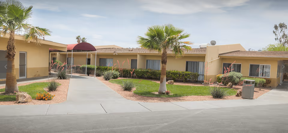 Front exterior of a single-story beige senior living facility with a central walkway, palm trees, landscaping, and a covered entrance.