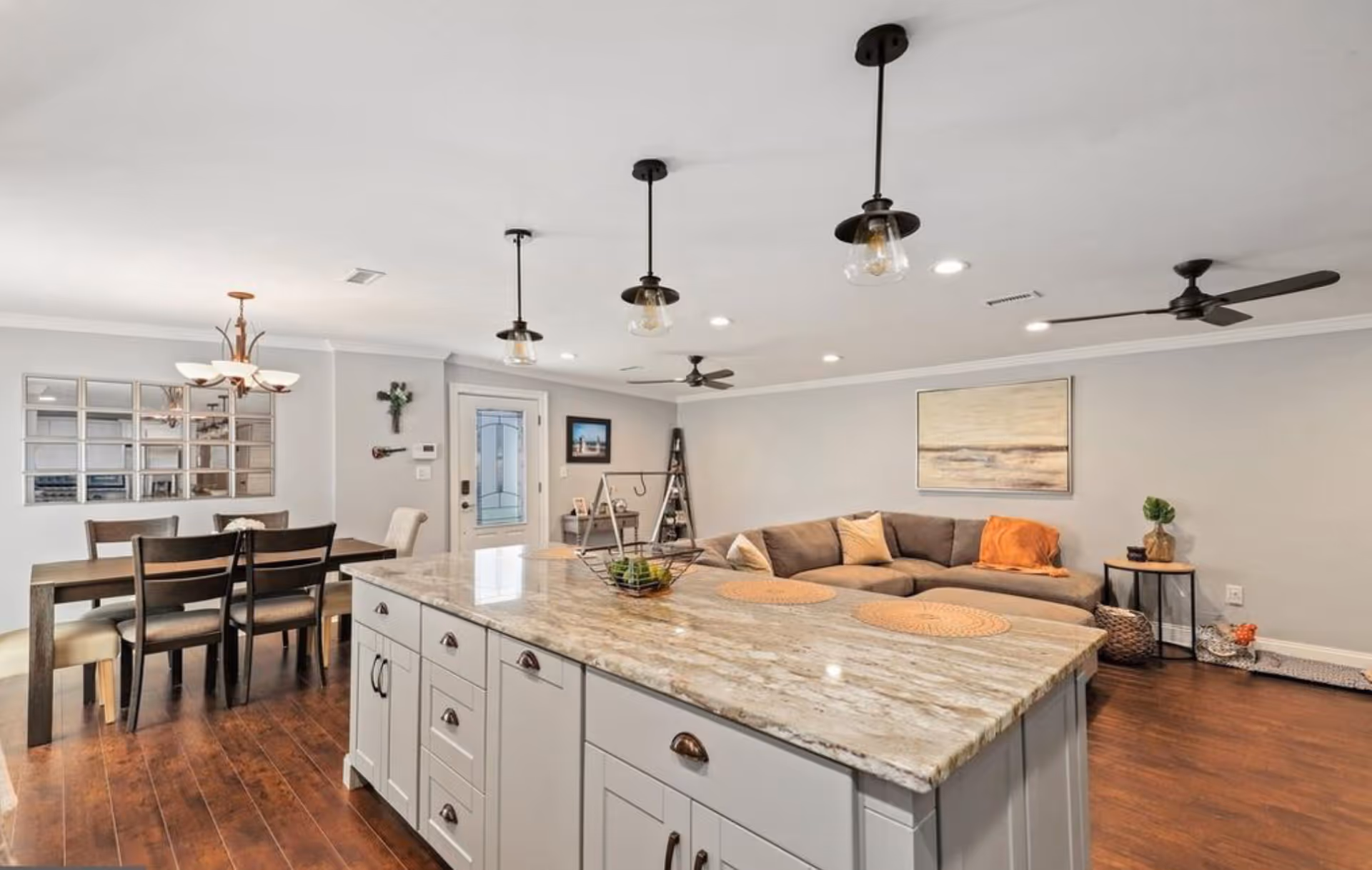 Open concept interior showing a kitchen island with a granite countertop in the foreground, a dining area with a wooden table and chairs to the left, and a living room with a large sectional sofa, wall art, and ceiling fans in the background.