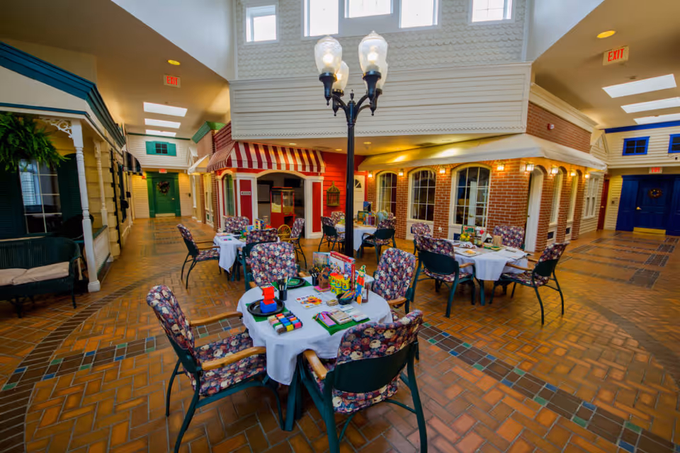 Indoor common area with round tables and floral-upholstered chairs set up for activities, surrounded by storefront-style facades and a central decorative streetlamp.