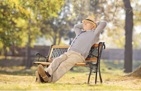 An elderly man wearing a hat and gray sweater is reclining and relaxing on a park bench outdoors with trees and sunlight in the background.