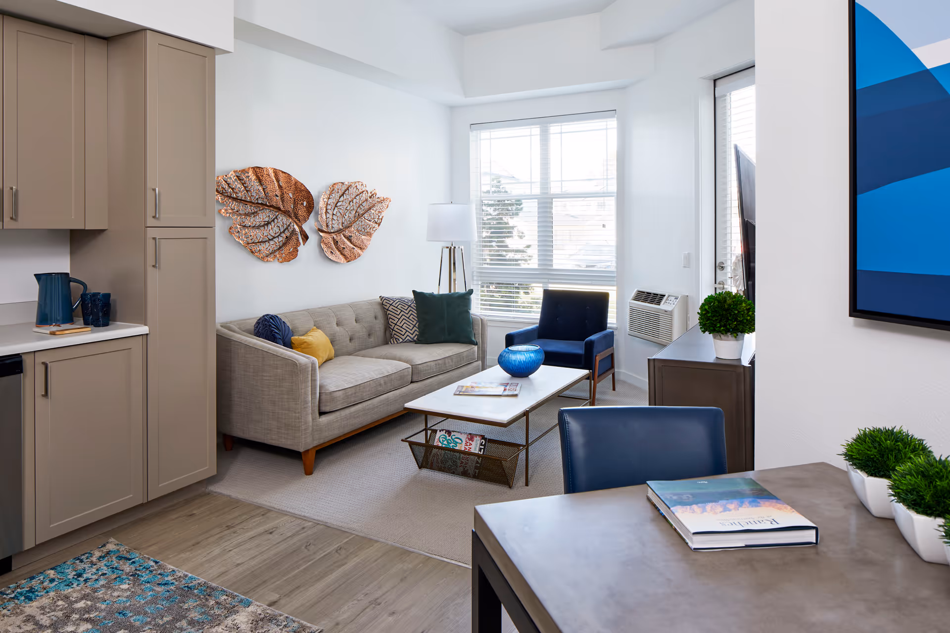 A bright and modern living room area with a beige sofa adorned with colorful pillows, a blue armchair, and a white coffee table with a blue decorative bowl and magazines. The room features large windows with blinds, a floor lamp, and wall art of two large metallic leaves. Adjacent to the living area is a kitchen with taupe cabinets and a countertop holding a blue pitcher and cups. A table with a book and small potted plants is also visible in the foreground.