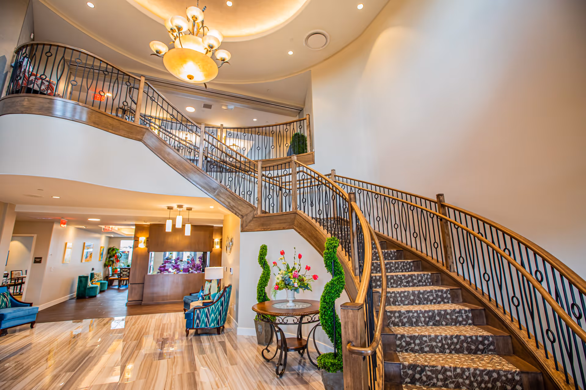 Grand curved wooden staircase in a senior living facility lobby with patterned carpet, chandelier, seating, and a floral table.