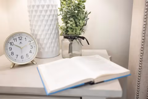 A bedside table with an open book, a round white clock showing 10:10, a white textured vase, and a small glass jar with green leafy stems.