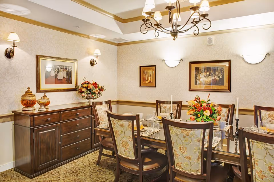 A well-lit dining room with a wooden dining table set for six people. The chairs have floral upholstery. There are two floral centerpieces on the table along with candles and glassware. The walls are decorated with framed paintings and wall sconces. A wooden sideboard with decorative jars and a flower arrangement is against one wall.