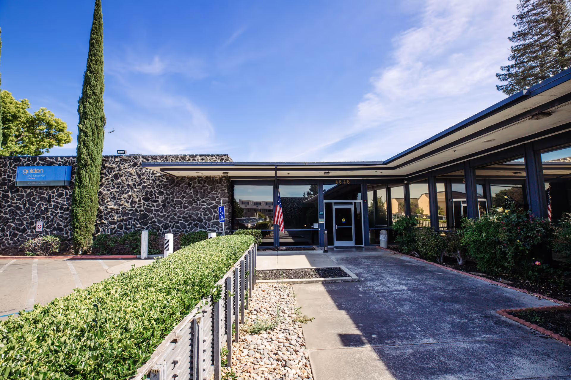 Exterior view of Stockton Nursing Center showing the entrance with glass doors, an American flag, a tall cypress tree, and a stone facade wall under a clear blue sky.