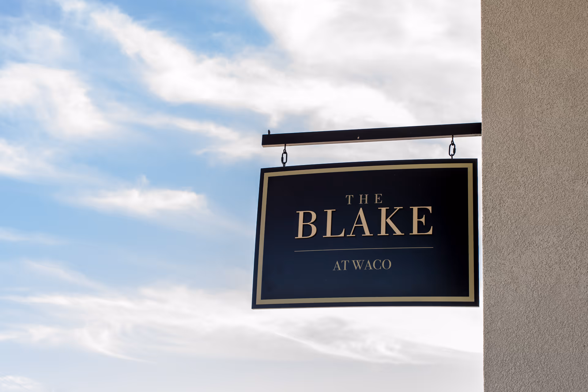 A black and gold hanging sign attached to a building wall with the text 'THE BLAKE AT WACO' against a partly cloudy blue sky background.