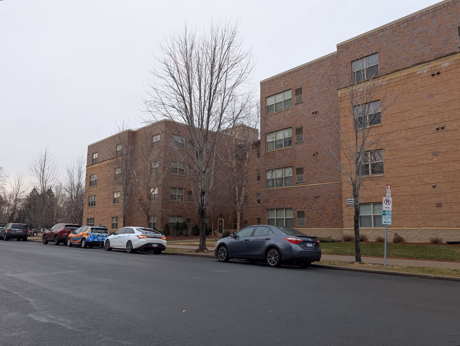 Street view of a multi-story brick residential building with parked cars and bare trees.