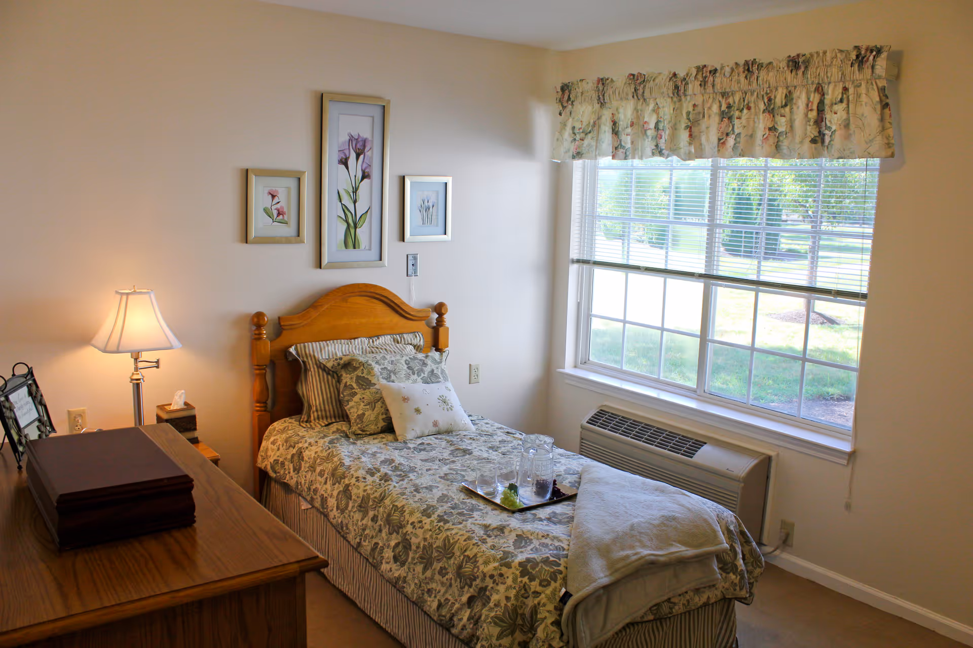 Sunny single bedroom with a wooden twin bed dressed in floral bedding, a bedside lamp and dresser, and a large window overlooking greenery.