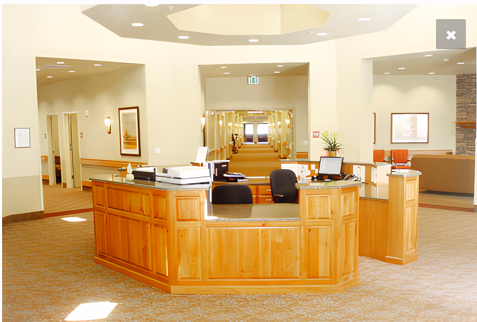 Wooden reception desk in a bright senior living facility lobby with chairs, a seating area to the right, and a long hallway visible behind it.