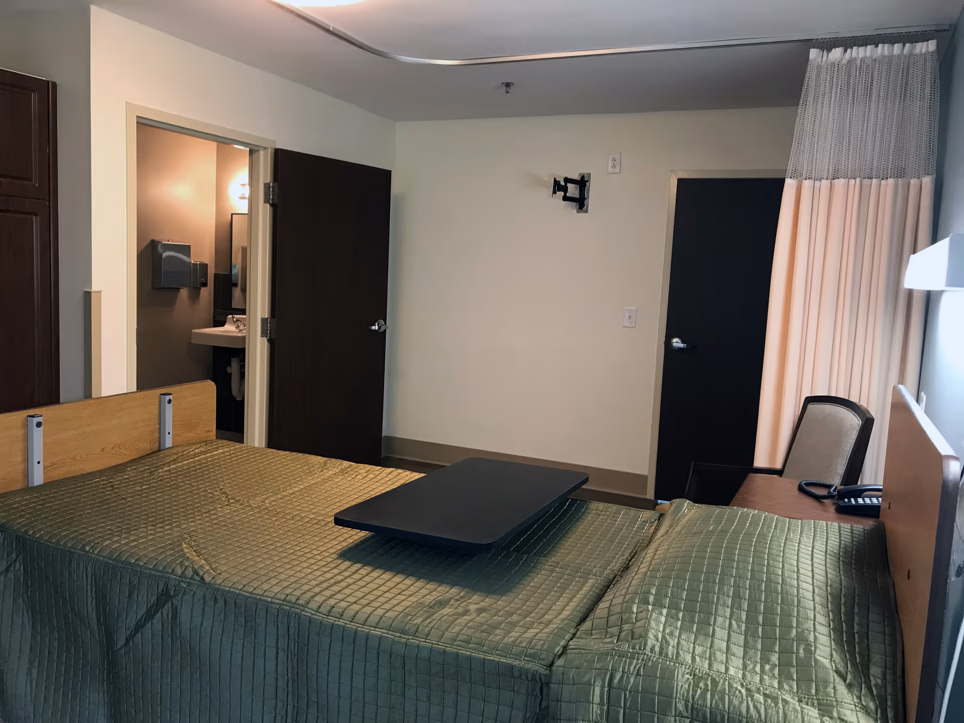 A simple bedroom in a senior living facility with a bed covered in a green quilted blanket and a black overbed table. There is a chair and a telephone on a small table next to the bed. The room has beige walls, a door leading to a bathroom with a sink and mirror visible, and another closed door. A curtain is partially drawn on the right side of the room.