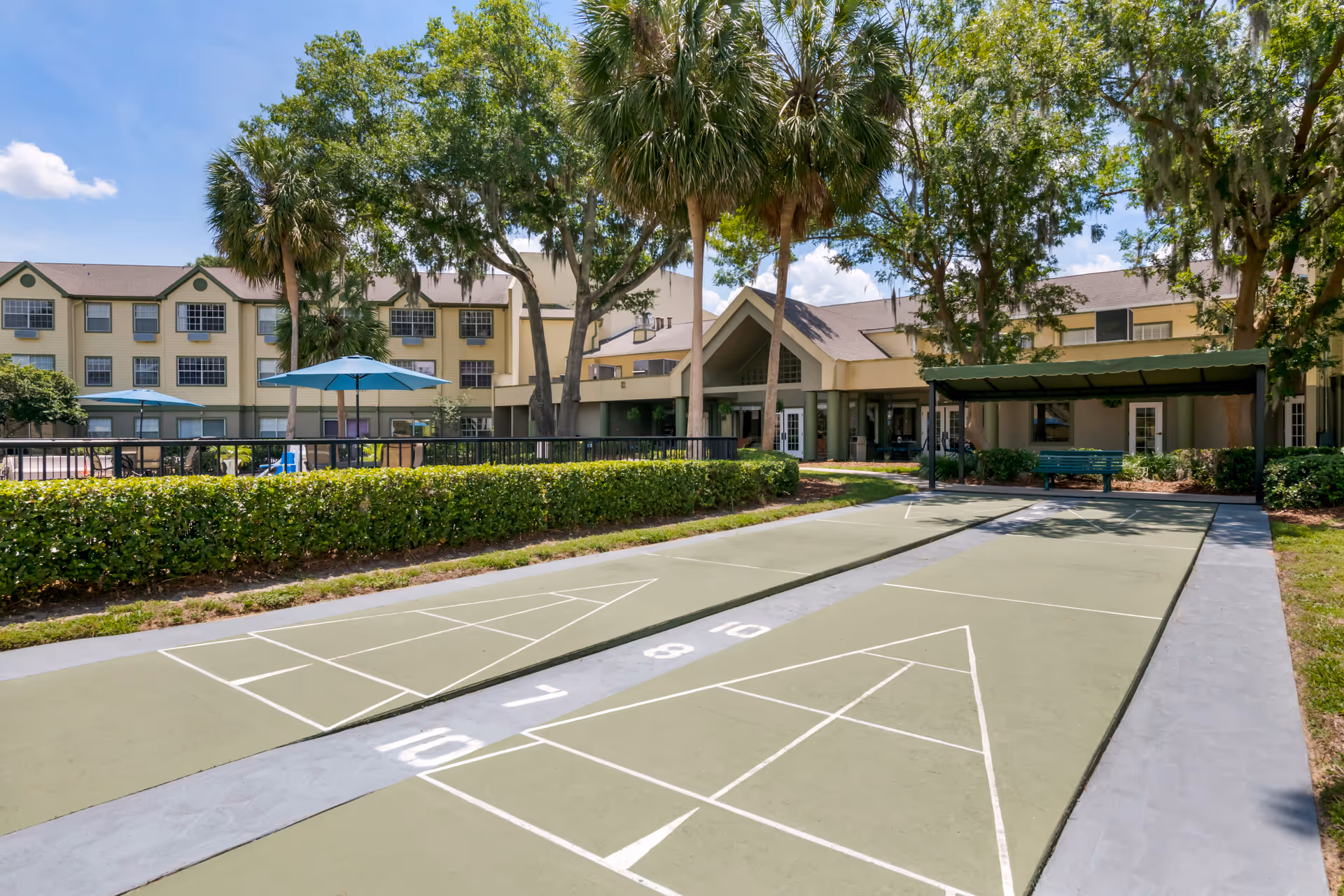 Outdoor shuffleboard courts with numbered markings in front of a senior living facility building. The area is surrounded by green hedges, palm trees, and other trees. There are blue umbrellas and seating areas near the building, which has a beige exterior and multiple windows under a clear blue sky.