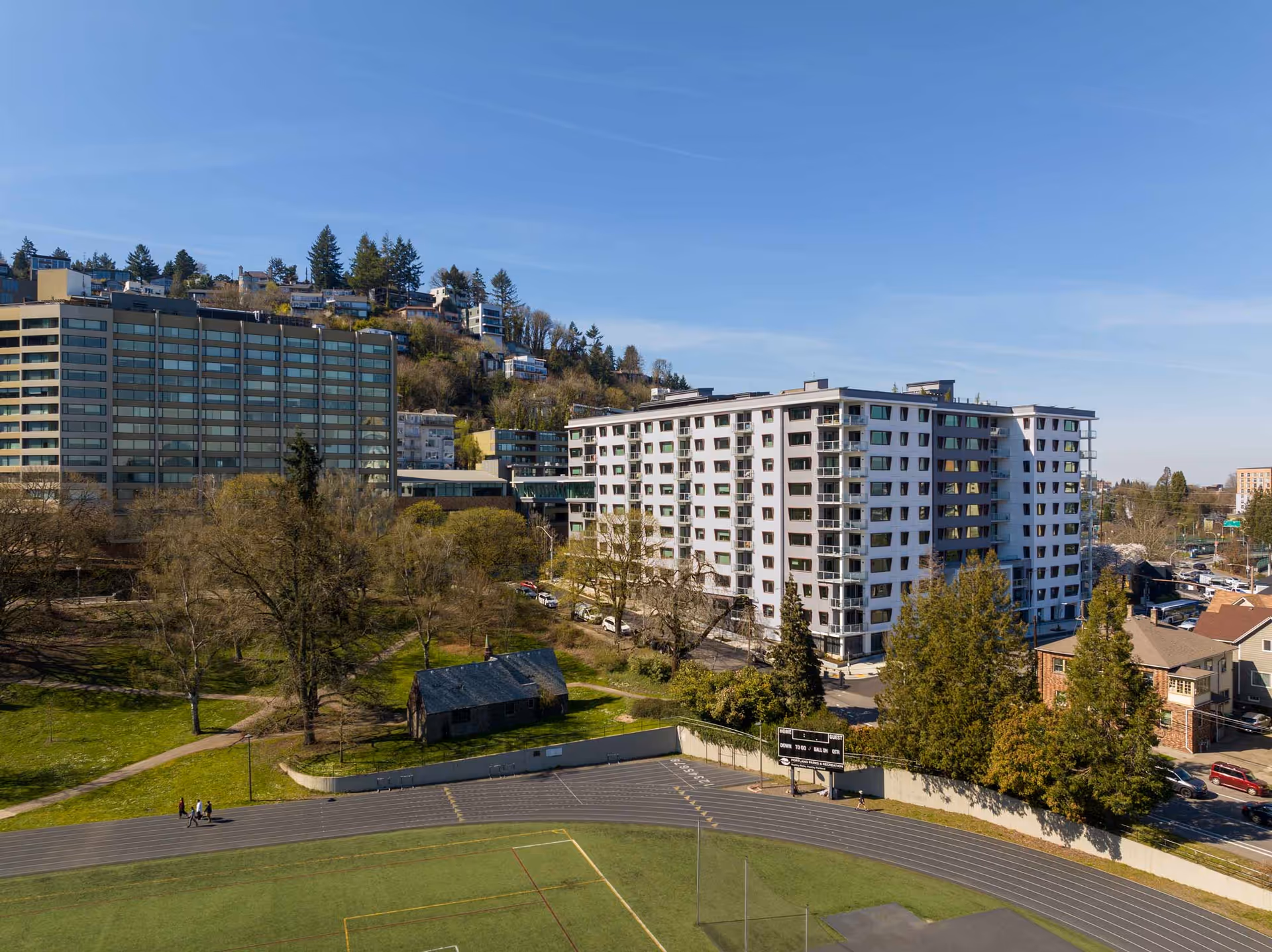 Aerial view of a multi-story residential building surrounded by trees and greenery, with a sports field and running track in the foreground. Additional buildings and houses are visible in the background under a clear blue sky.