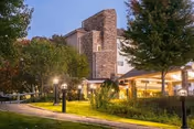 Evening exterior view of a senior living facility named Brookhaven At Lexington, showing a well-lit building with stone and stucco facade, surrounded by trees and landscaped greenery along a paved walkway.