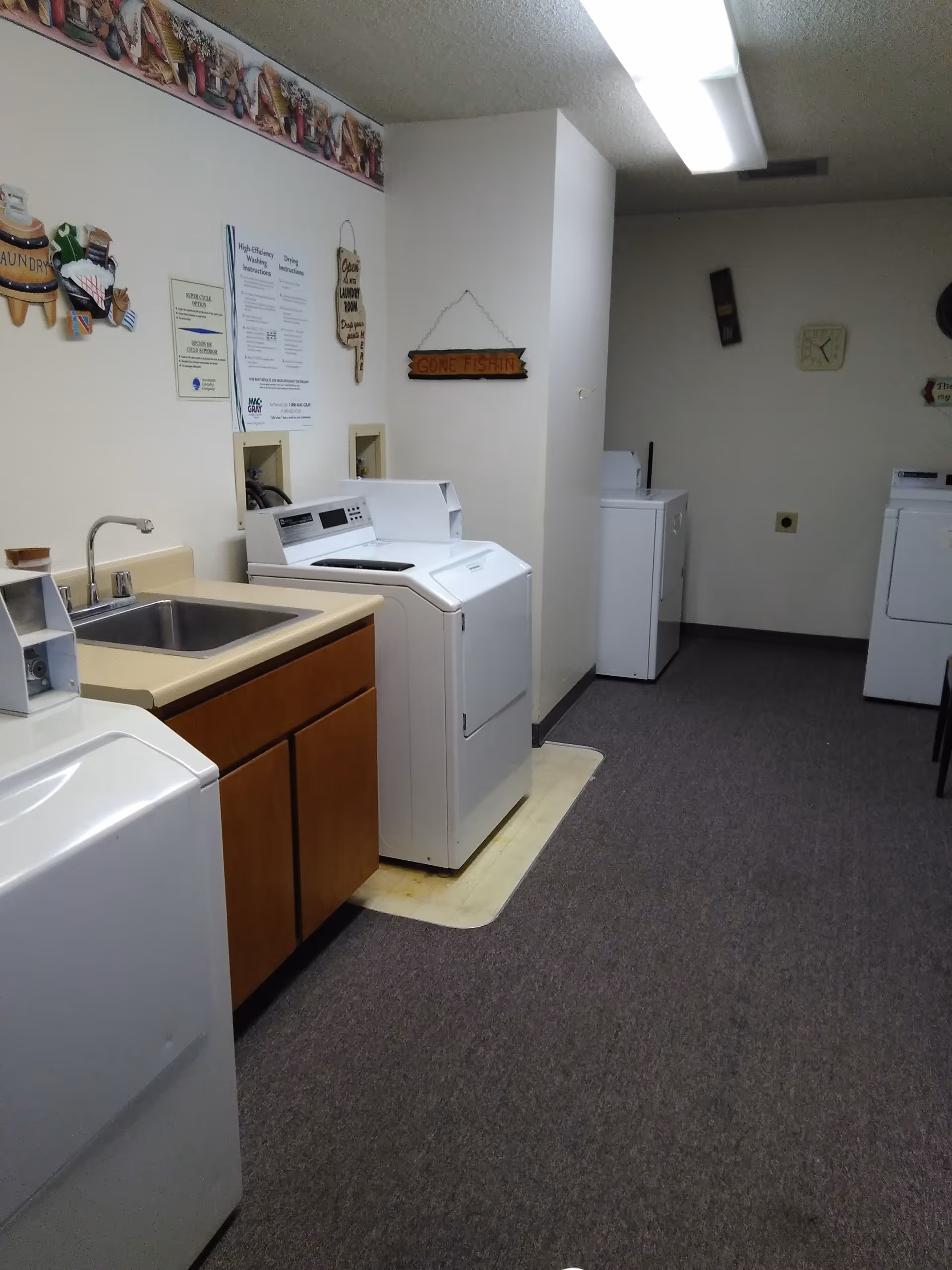 Laundry room with multiple white washing machines and dryers, a sink with a faucet, wooden cabinets below the sink, and various decorative signs on the walls including one that says 'Gone Fishin'. The room has carpeted flooring and fluorescent ceiling lighting.