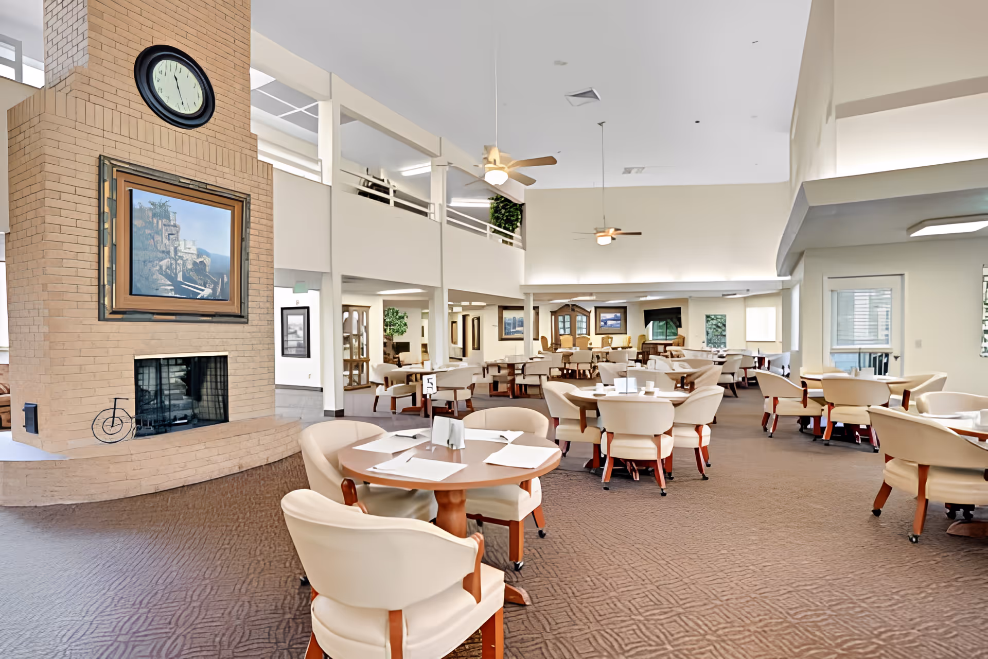 Spacious dining room in a senior living facility with multiple round tables and cream-colored chairs. A large brick fireplace with a clock and a framed painting is visible on the left side. The room has high ceilings with ceiling fans and soft lighting, and there are various framed pictures on the walls.