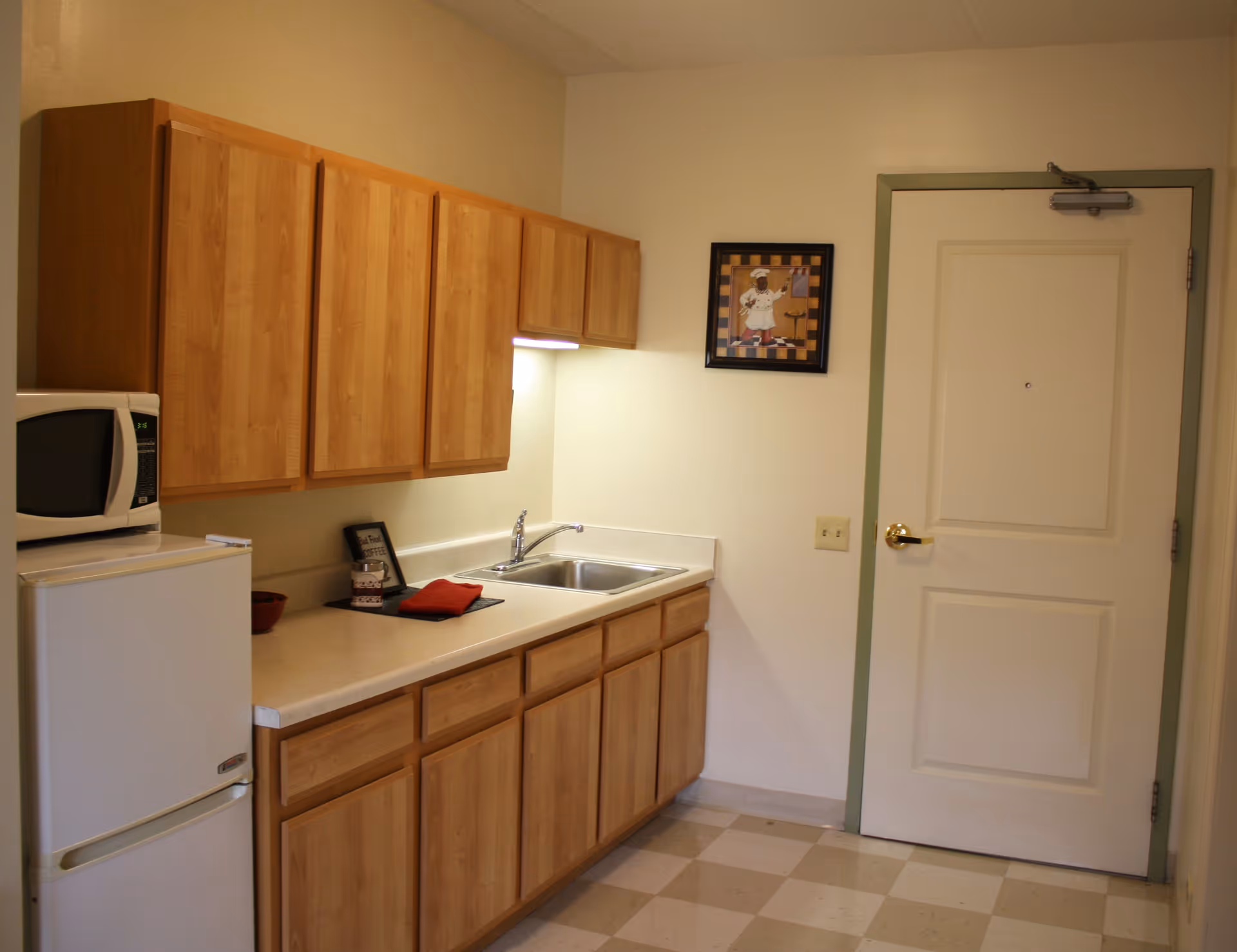 A small kitchen area with light wood cabinets above and below a beige countertop. There is a white microwave on top of a white mini refrigerator on the left side. A stainless steel sink is installed in the countertop with a faucet. A framed picture of a chef hangs on the wall next to a white door with a green trim. The floor has a checkered pattern with beige and white tiles.