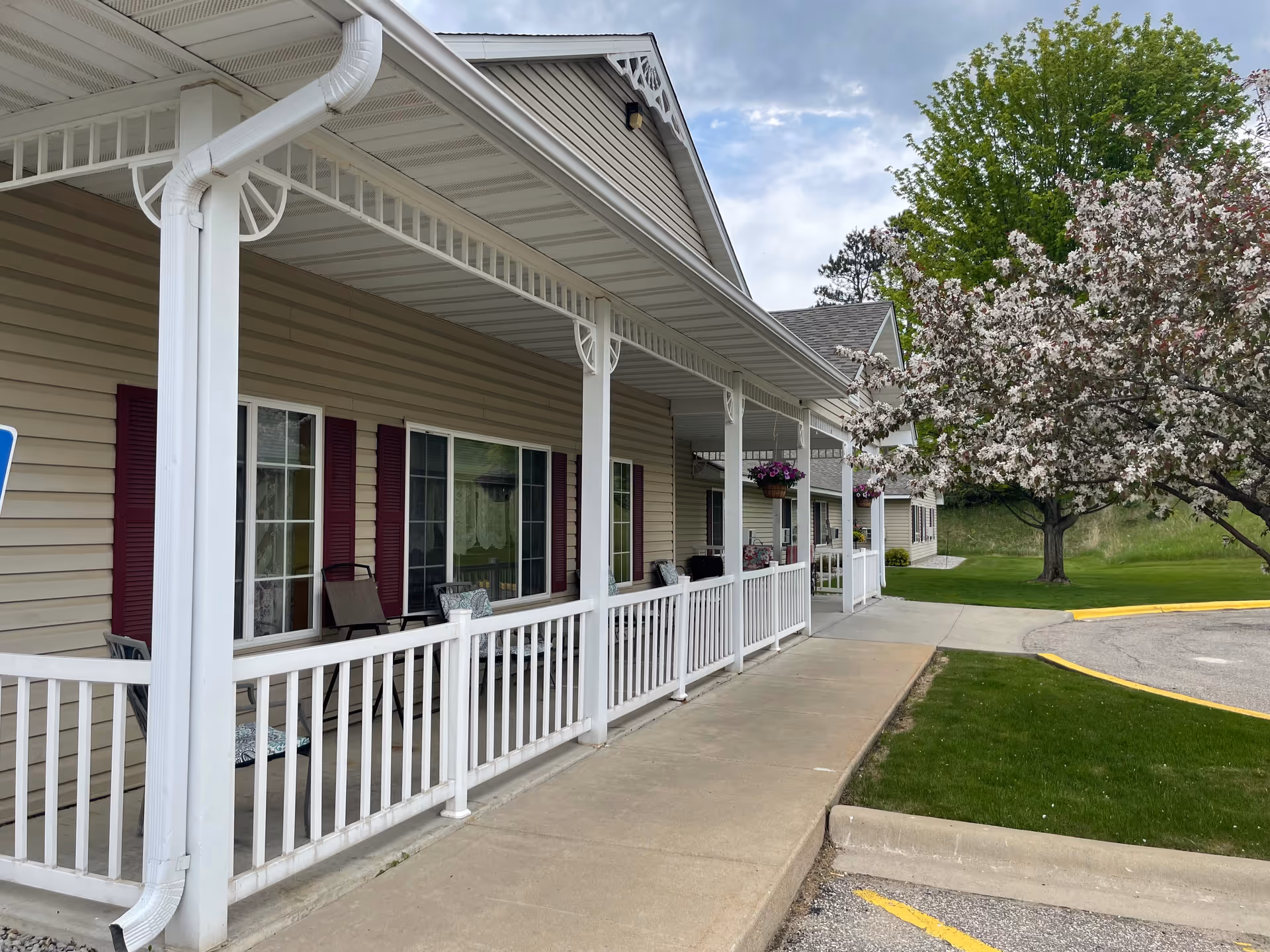 Covered front porch and entrance of a beige assisted living building with white railing, chairs, hanging flowers, and a blooming tree nearby.