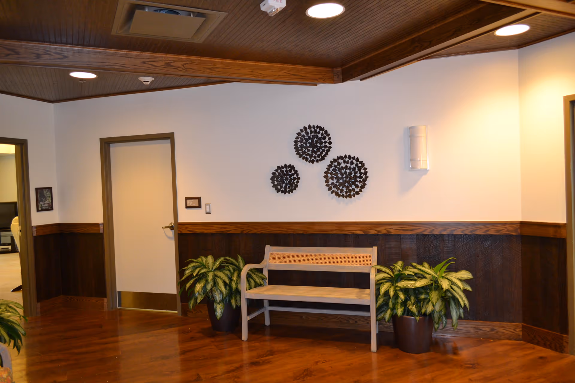 Interior hallway area with wooden floor and wood-paneled lower walls. A light-colored wooden bench is placed against the wall, flanked by two potted green plants. Three decorative black circular wall hangings are mounted above the bench. There are two closed doors and one open doorway visible, with warm lighting from ceiling fixtures.