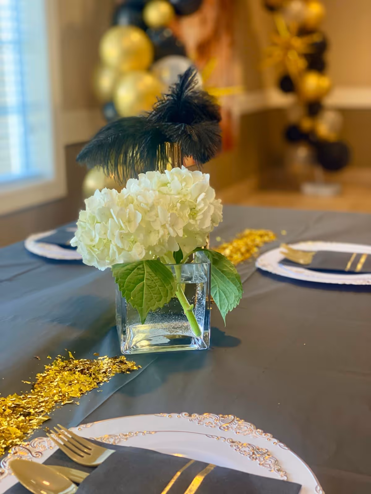 A close-up of a dining table set with white plates with gold trim, black napkins with gold stripes, and gold-colored cutlery. A square glass vase with white hydrangea flowers and black feathers serves as the centerpiece. Gold confetti is scattered on the dark tablecloth. In the background, there are black and gold balloons.