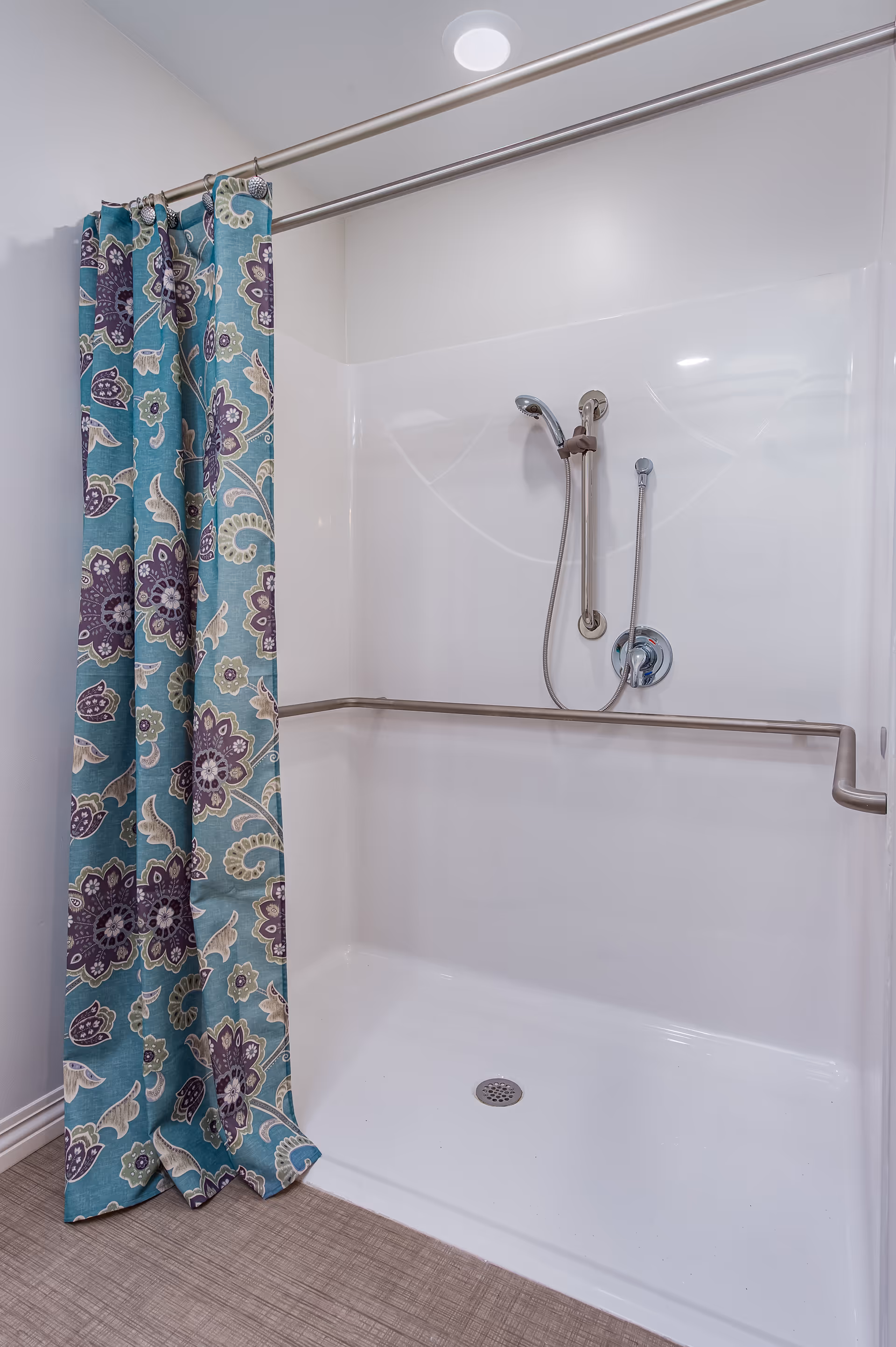 Walk-in shower with a teal floral shower curtain, handheld showerhead, and stainless grab bars.
