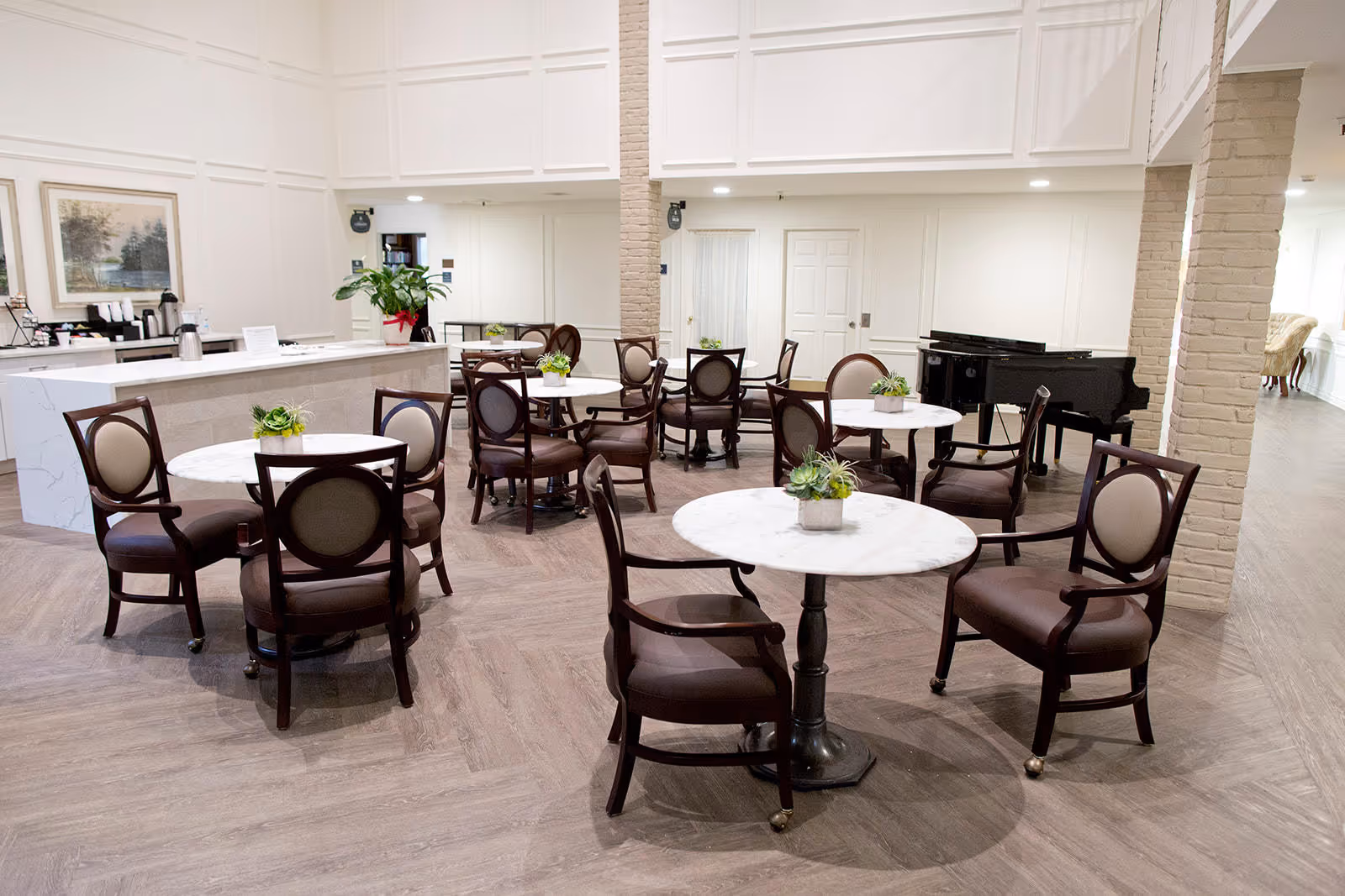 A bright and spacious dining area with several round marble-top tables surrounded by dark wooden chairs with cushioned seats and backs. Each table has a small green plant centerpiece. In the background, there is a white counter with coffee dispensers and a large plant. A black grand piano is positioned near the back wall, and the room features light-colored walls and wood-patterned flooring.